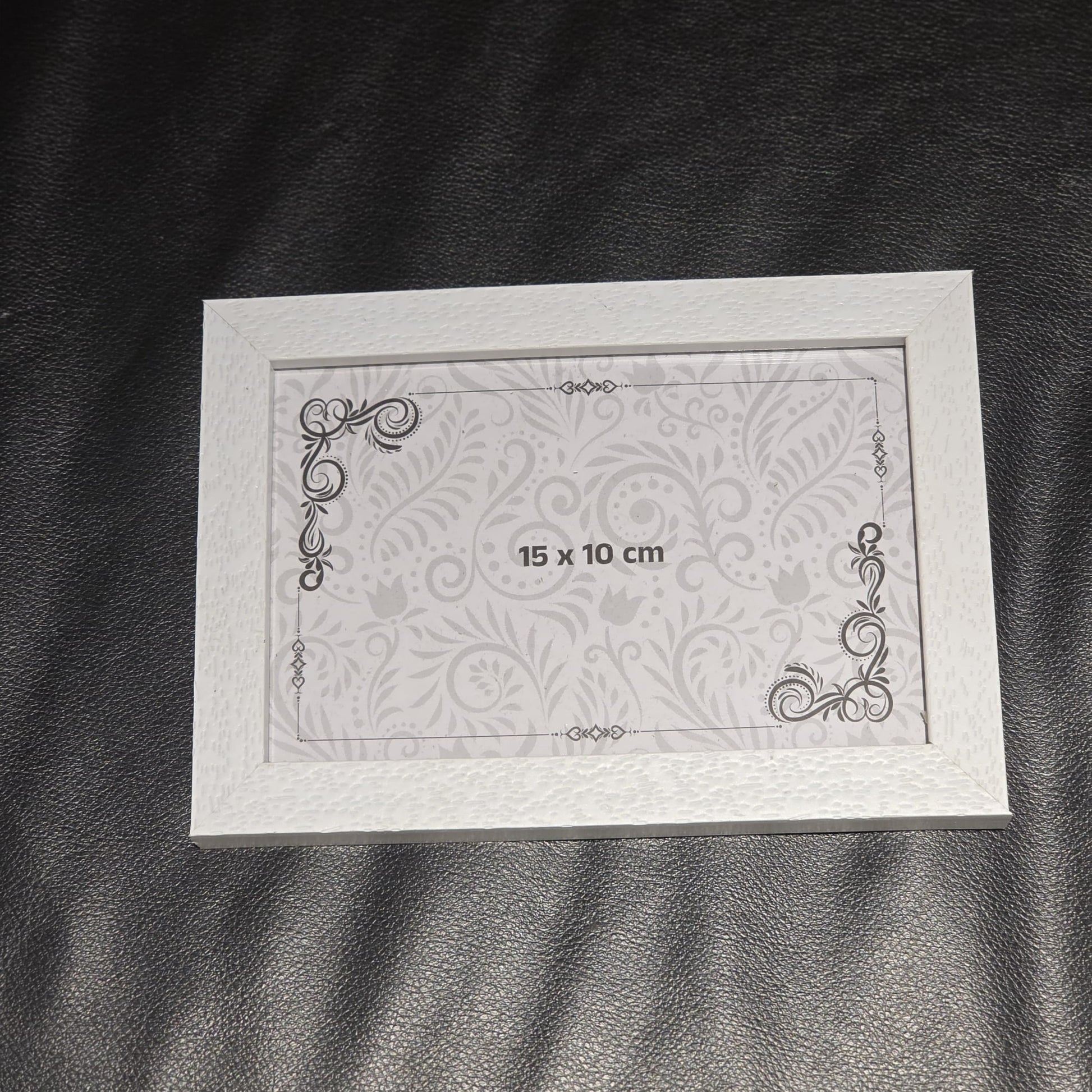 A 15 x 10 cm white picture frame with a subtle damask pattern and decorative scroll corners, displayed on a black textured surface.