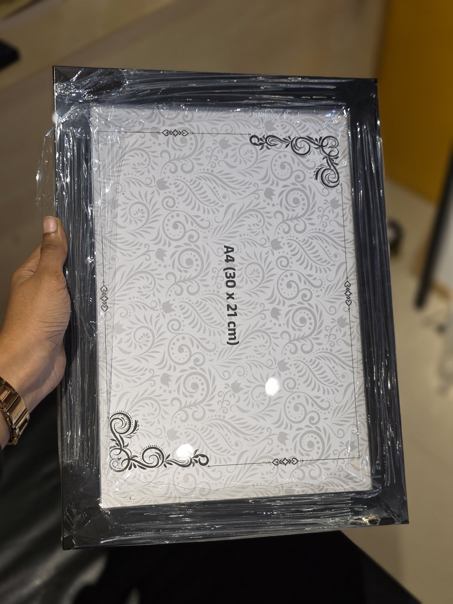 A hand holding a shrink-wrapped A4 size picture frame with a black ornate border and a white patterned insert, shown in a real indoor setting.