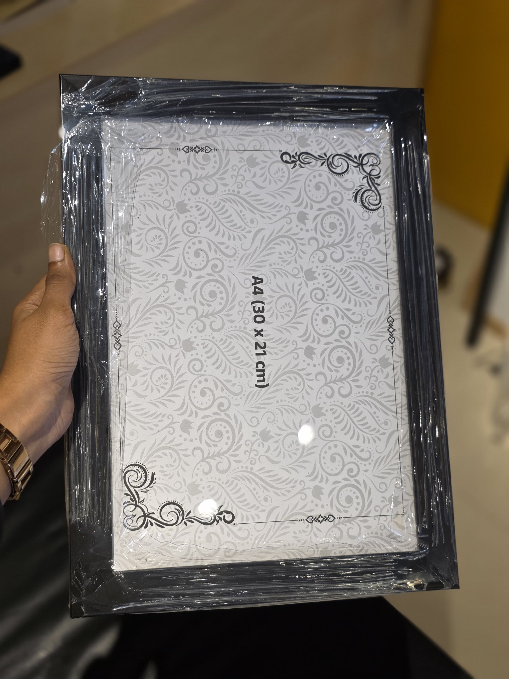 A hand holding a shrink-wrapped A4 size picture frame with a black ornate border and a white patterned insert, shown in a real indoor setting.