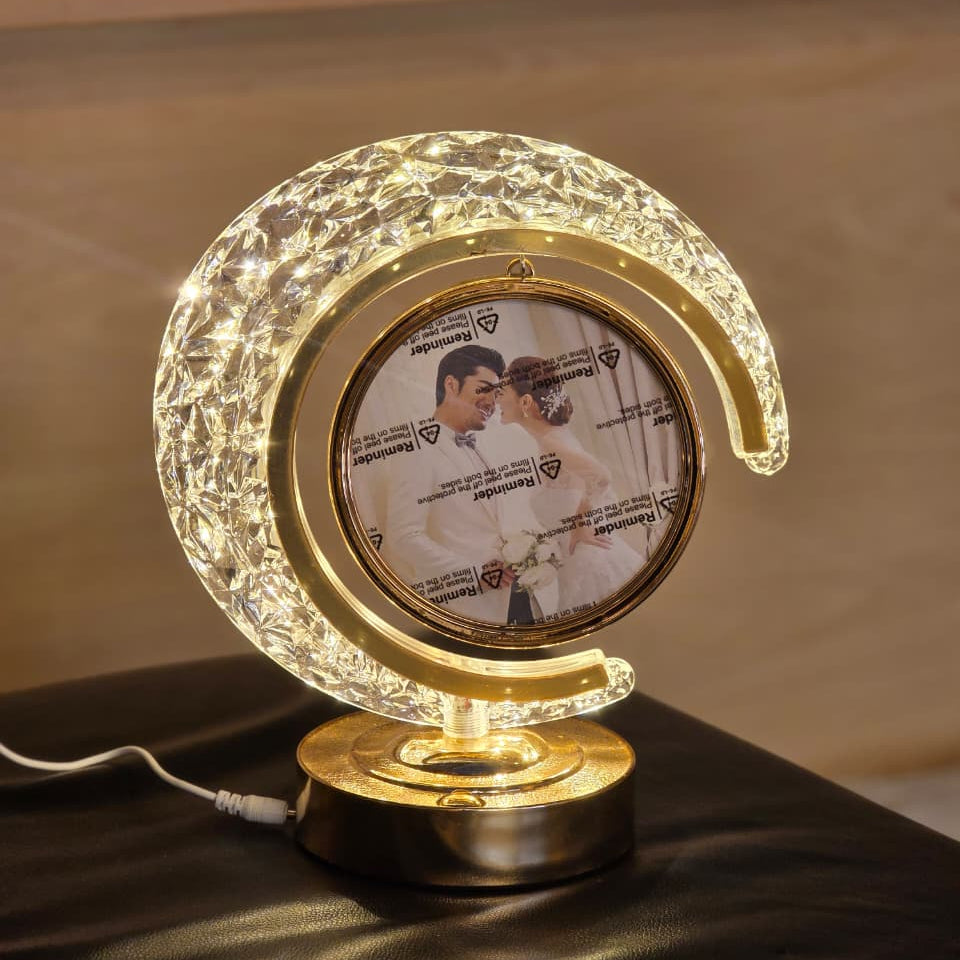 A crescent moon-shaped LED night light with a gold-toned frame and base, featuring a custom photo insert in the center, displayed on a dark surface with a blurred indoor background.