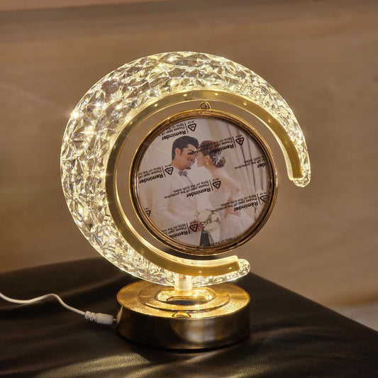 A crescent moon-shaped LED night light with a gold-toned frame and base, featuring a custom photo insert in the center, displayed on a dark surface with a blurred indoor background.