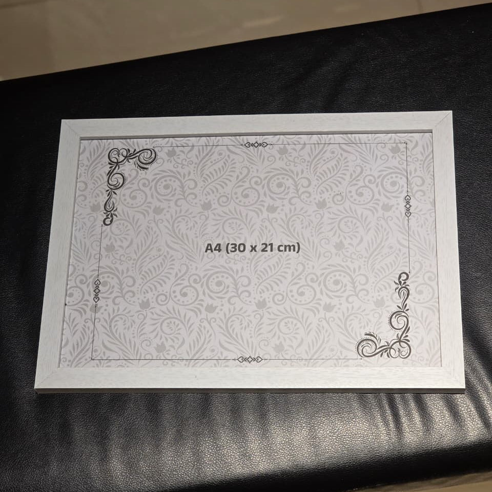 A square white A4 photo frame insert with a subtle floral damask pattern and decorative corner flourishes, labeled 'A4 (30 x 21 cm)', resting on a black leather surface.