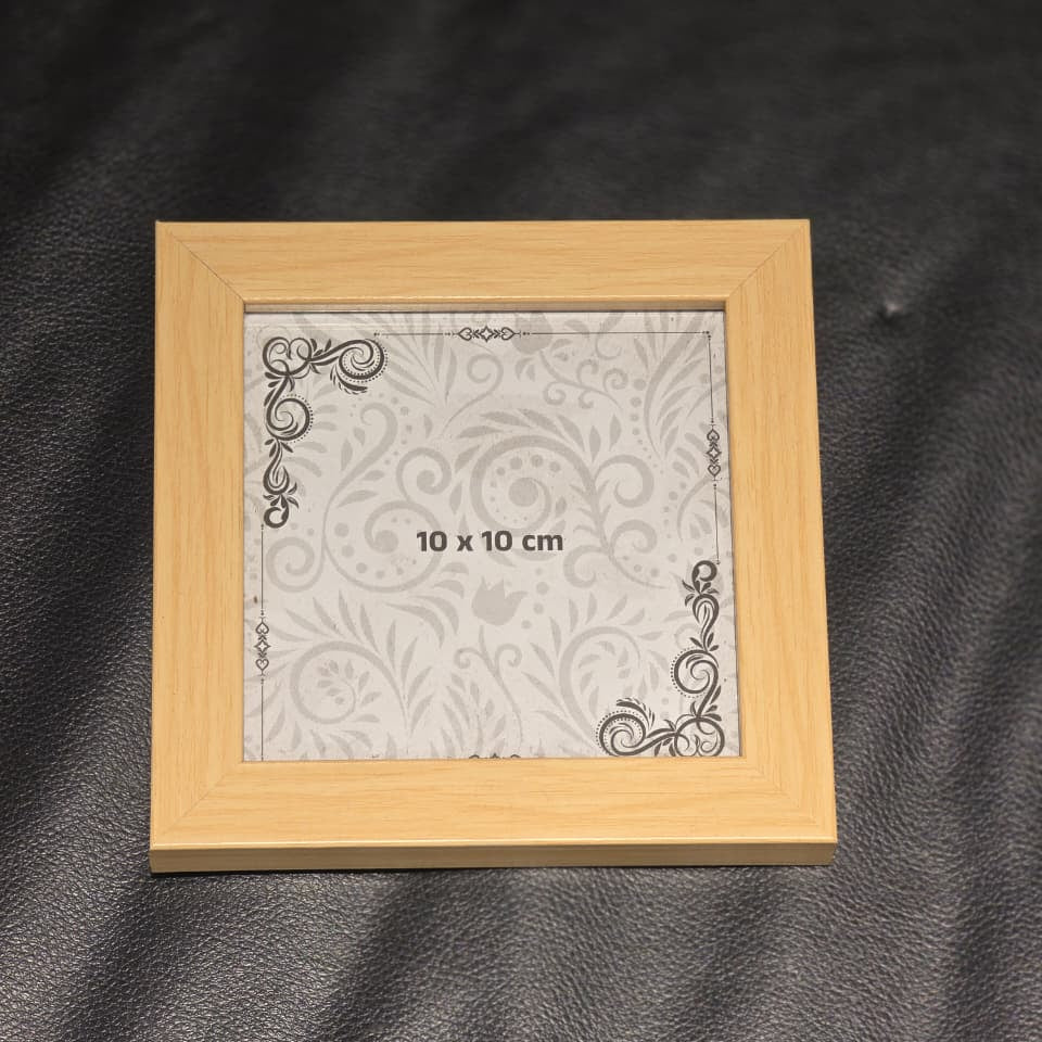 A small square picture frame made of light natural wood, containing a white patterned insert with '10 x 10 cm' printed in the center, placed on a dark textured surface.