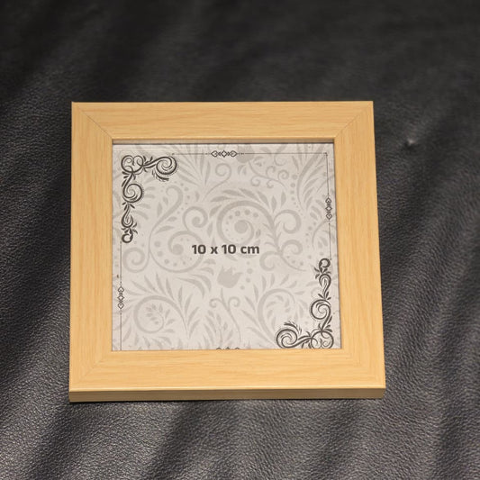 A small square picture frame made of light natural wood, containing a white patterned insert with '10 x 10 cm' printed in the center, placed on a dark textured surface.