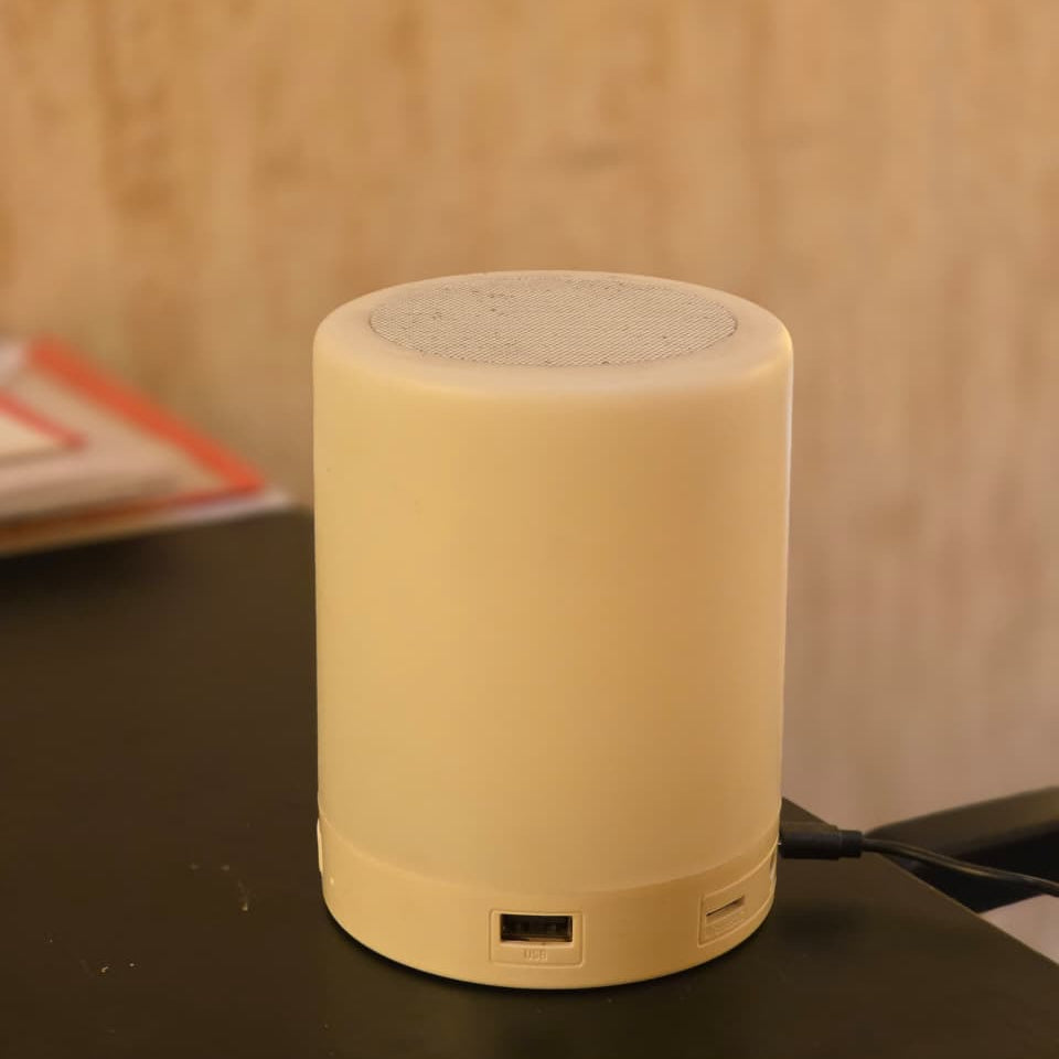 A small cylindrical cream-colored portable speaker with a textured top grille, sitting on a dark tabletop against a warm, blurred background.
