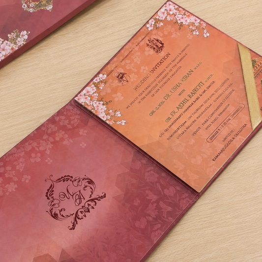 A traditional Premium wedding invitation suite displayed on a light wood surface, featuring a deep pink envelope with gold floral details and a peach-colored invitation card decorated with cherry blossoms.