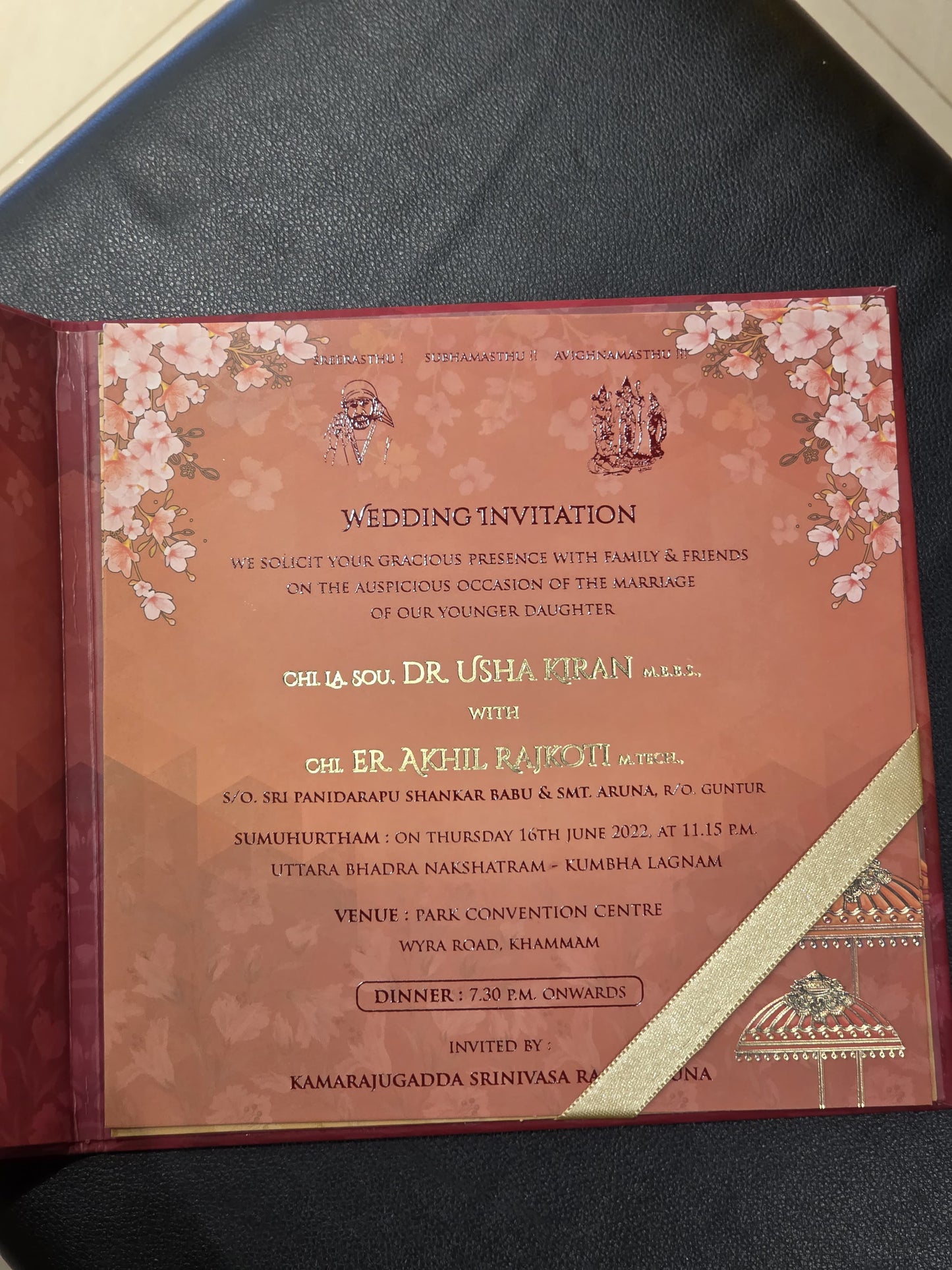 A printed wedding invitation card with a peach and pink floral design, placed on a dark textured surface. The card includes decorative cherry blossom illustrations, ornate borders, and a diagonal gold ribbon detail.