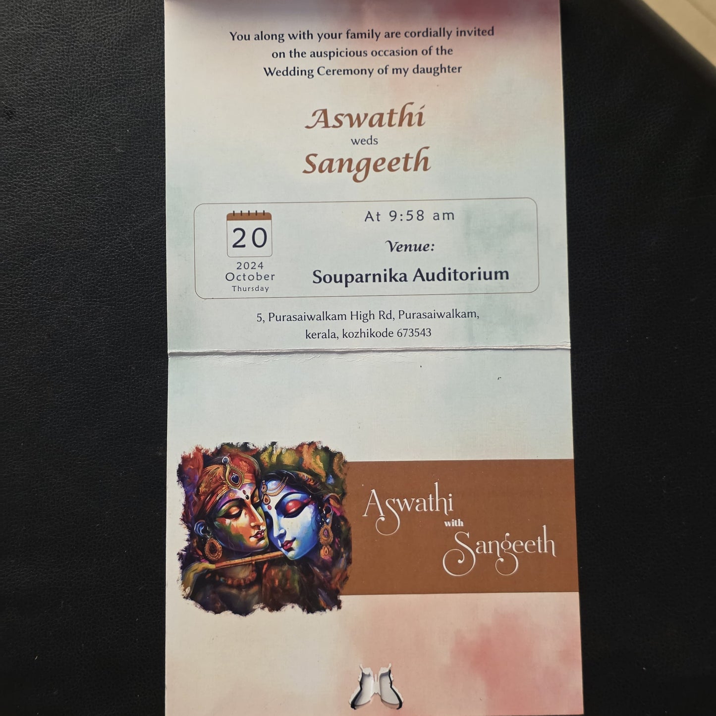 A wedding invitation card for 'Aswathi with Sangeeth' featuring a brown and pink design,  and event details, photographed on a dark textured surface.