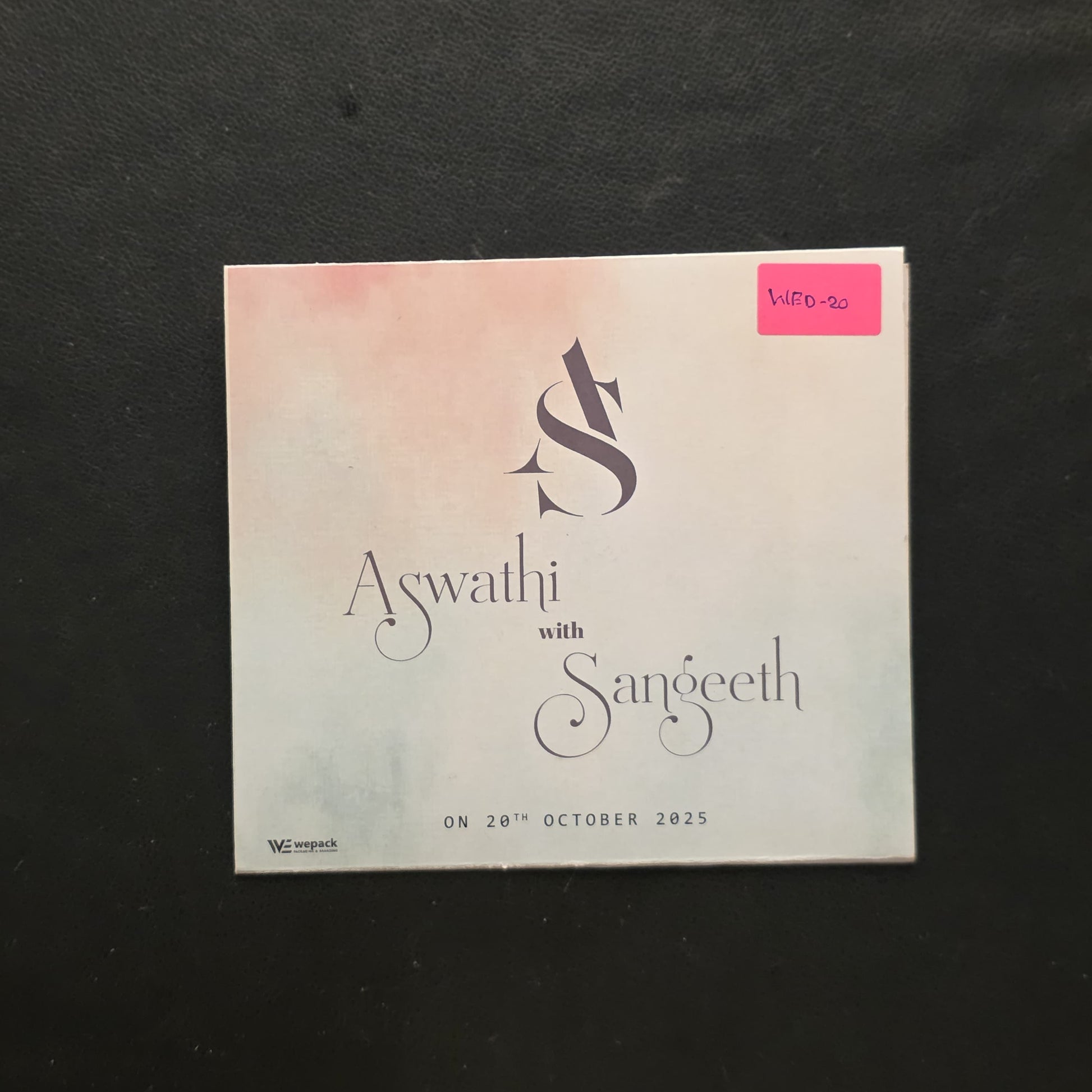 A square printed card or ticket with the text 'Aswathi with Sangeeth' and a stylized 'AS' logo, placed on a dark fabric background. The card has a soft pink-to-white gradient, a small pink price sticker in the top right, and event text at the bottom.