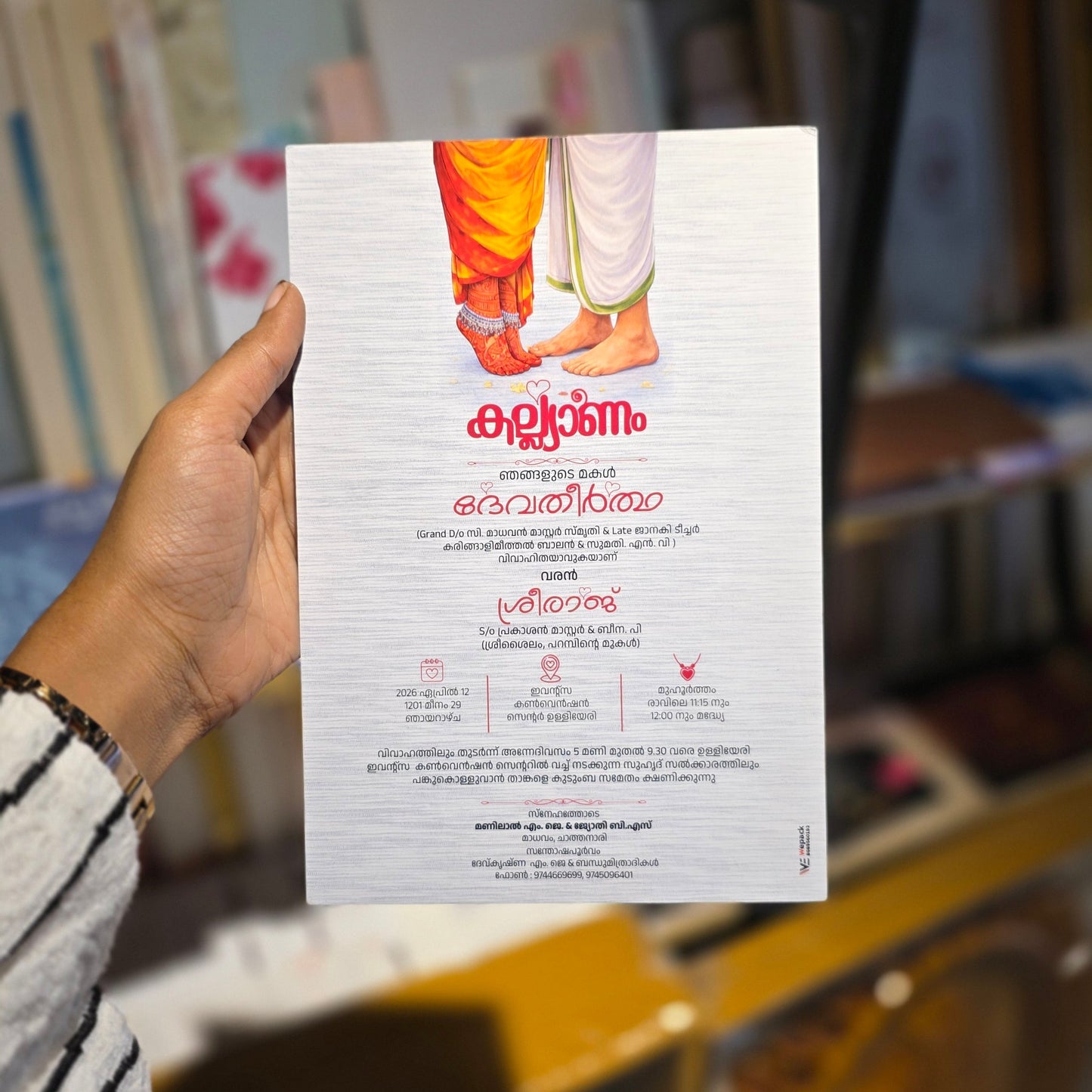A hand holding a printed wedding  card with malayalam text and a small photo of a foot, photographed in a real indoor environment with shelves and objects in the background.