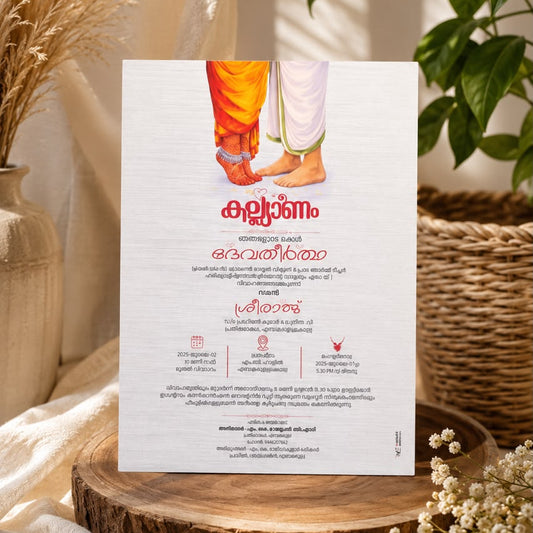 A vertical invitation card with malayalamt ext and an illustration of feet in orange and white dhotis, styled on a wooden slice with dried flowers and a potted plant in a warm indoor setting.