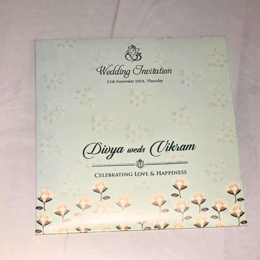 A square wedding invitation card with a pale mint green background, delicate floral illustrations, and elegant black text reading 'Wedding Invitation' and 'Divya weds Vikram' on a white fabric surface.